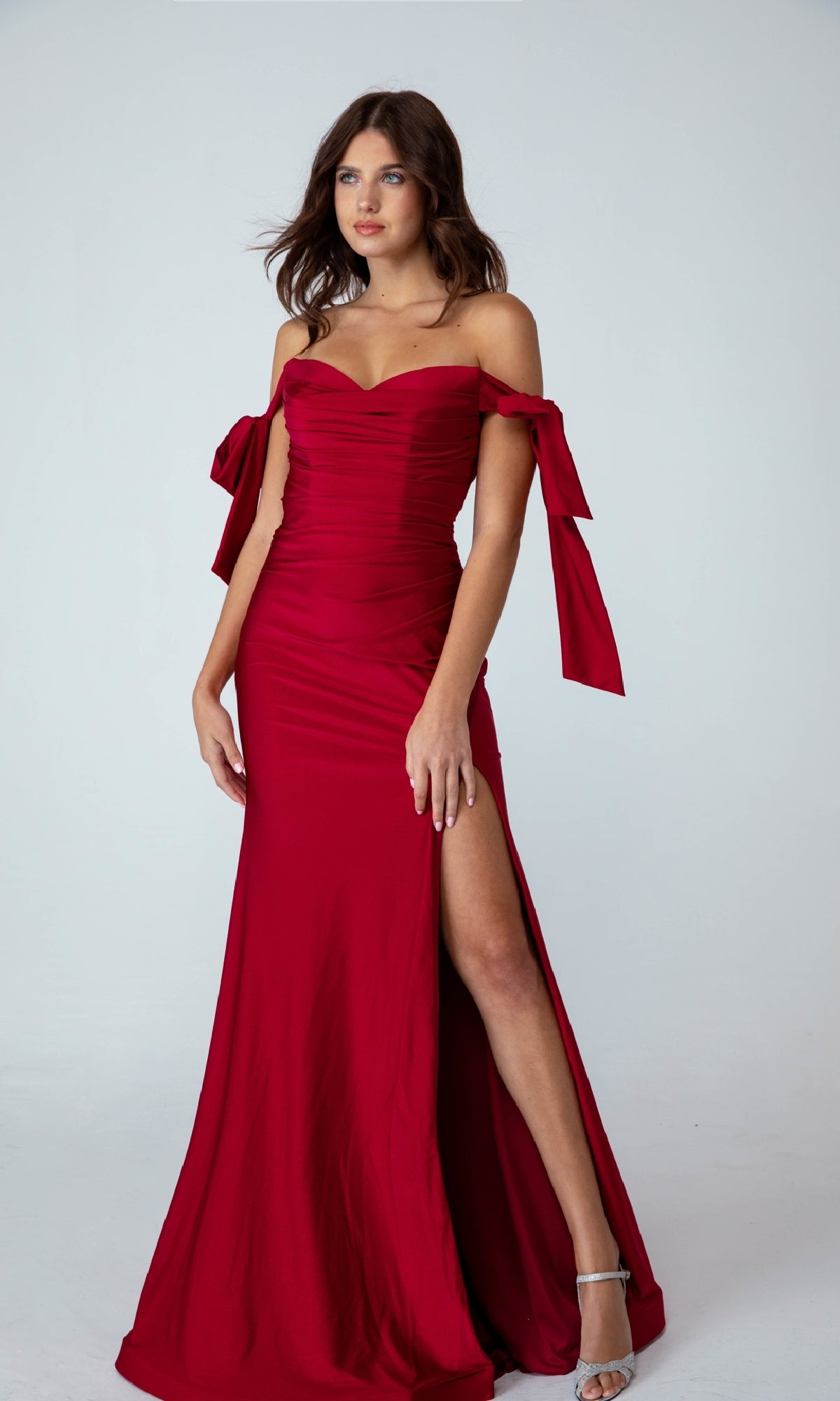 Woman in the Shoulder-Ties Long Jersey Prom Dress: Eureka 9181—an off-the-shoulder gown with a ruched bodice, large sleeve bows, thigh-high slit, and silver heels—stands before a plain background. Elegant and affordable.